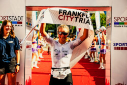 28.07.24: Triple für Sarah Schönfelder. Sie gewinnt die Mitteldistanz des Frankfurt City Triathlon zum dritten Mal. Im Ziel mit Siegerbanner